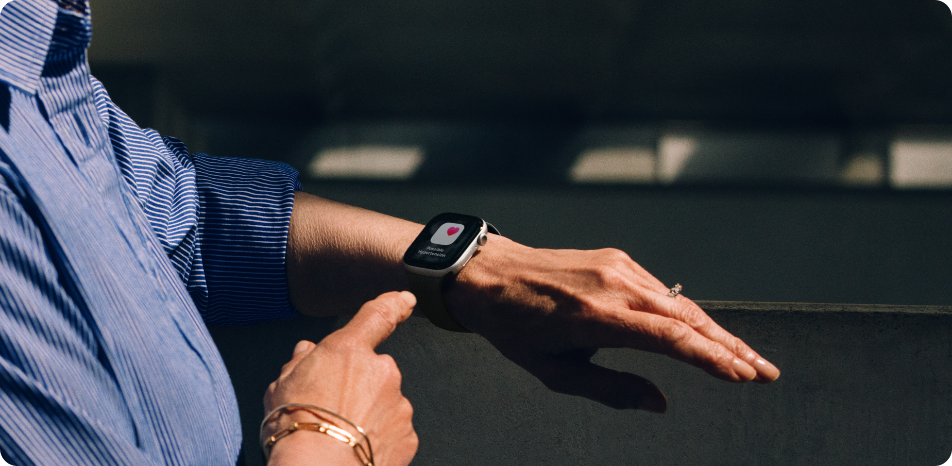 Eine Person, die ein blau gestreiftes Hemd trägt, interagiert mit einer Smartwatch an ihrem linken Handgelenk, die ein Aufnahmesymbol auf dem Bildschirm anzeigt. Die Szene ist gut beleuchtet, wobei der Hintergrund unscharf ist.