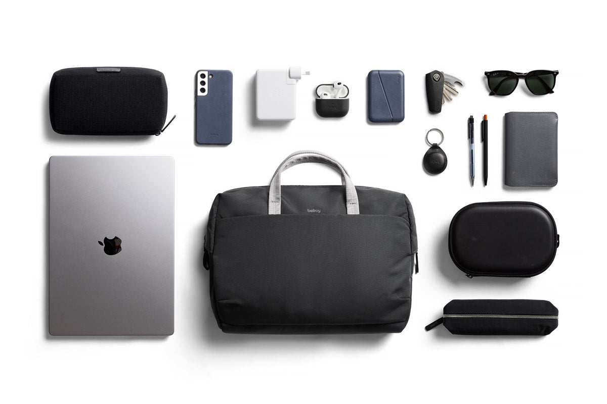Flat-Lay-Aufnahme einer Bellroy Via Work Bag (Tech Briefcase) in der Farbe „Slate“, umgeben von Zubehör: Laptop, Handy, Ladegeräte, AirPods, Stifte, Notizbuch, Sonnenbrille, Schlüssel, kleine Etuis, ein Beutel und ein tragbarer Lautsprecher – alles ordentlich auf einem weißen Hintergrund angeordnet.