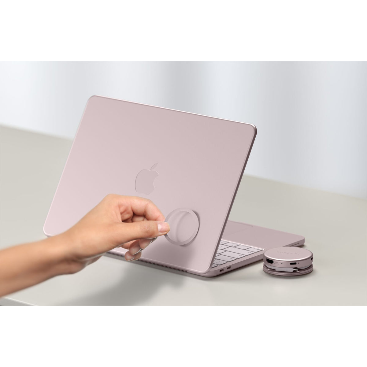 Eine Hand befestigt ein rundes, flaches Zubehörteil an der Rückseite eines rosa Apple-Laptops auf einem weißen Schreibtisch und unterstreicht damit die kompakte Bauweise Ihres Setups. Daneben befinden sich eine kabellose Tastatur und der Satechi OntheGo 5-in-1-Multiport-Adapter (Blush) von Satechi, die Ihrem Arbeitsplatz sowohl Stil als auch Funktionalität verleihen.