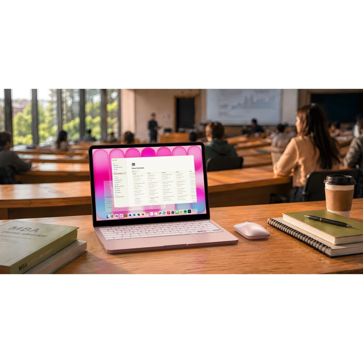 Auf einem Schreibtisch in einem Klassenzimmer steht ein Laptop mit einem Bildschirm in Rosa. Daneben liegen Notizbücher, eine Kaffeetasse, ein Stift und die „Satechi Slim EX Wireless Mouse (Blush)“ von Satechi. Die Schüler sitzen dem Lehrer an der Vorderseite des Raums gegenüber.