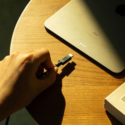 Eine Hand hält ein „Native Union Belt Cable XL“ USB-C-zu-USB-C-Kabel (3 m) in der Farbe „Cosmos“ neben einem iPad auf einem Holztisch, während Sonnenlicht Schatten über die Szene wirft.