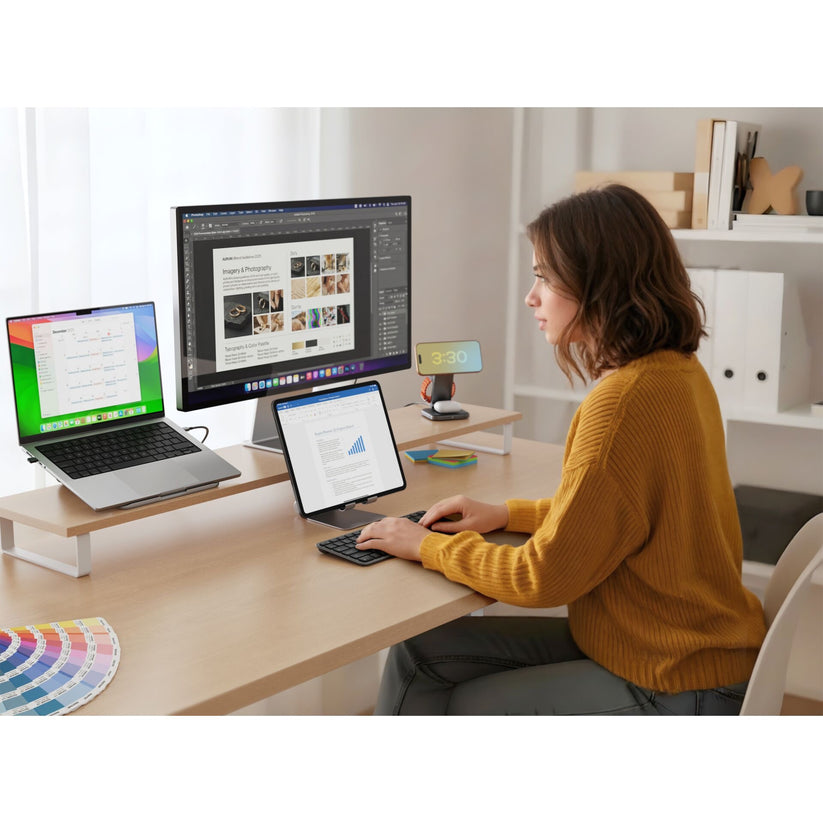 Eine Frau in einem gelben Pullover arbeitet an einem Schreibtisch mit Laptop, Tablet, einer kabellosen Tastatur – der „Satechi Slim EX1 Wireless Keyboard Space Black DE“ von Satechi mit deutschem Layout – sowie einem großen Monitor, auf dem Grafiken und Dokumente angezeigt werden. Außerdem sind im Raum eine Farbpalette, eine Uhr und Regale mit Boxen zu sehen.