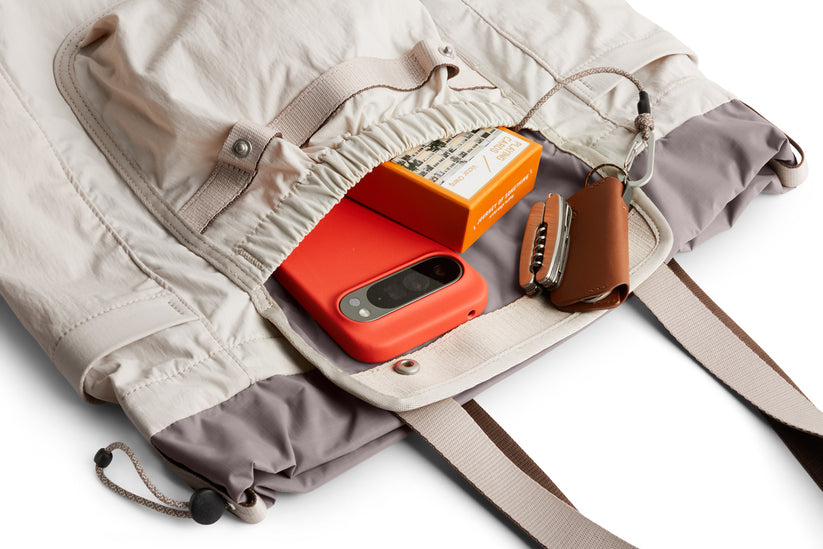Die „Bellroy Cinch Tote – Oat“ von Bellroy ist eine hellbeige-graue Tasche, deren Vordertasche offen ist und in der ein rotes Smartphone, eine orangefarbene Schachtel, ein brauner Schlüsselanhänger aus Leder und ein kompaktes Multitool ordentlich verstaut sind. Diese wasserabweisende Tragetasche verfügt über große Taschen für zusätzlichen Stauraum und sorgt dafür, dass Ihre wichtigsten Utensilien unterwegs gut organisiert und geschützt sind.