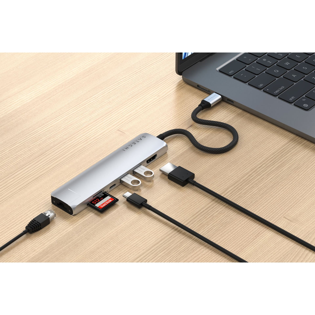 Gümüş renkli, Ethernet özellikli bir Satechi USB-C 7′si 1 arada İnce Çoklu Bağlantı Noktalı Adaptör, bir dizüstü bilgisayara bağlıdır ve USB kabloları, bir SD kart, bir microSD kart ve bir Ethernet kablosu dahil olmak üzere çeşitli cihazları barındırır; tüm bunlar açık renkli ahşap bir yüzey üzerinde düzenli bir şekilde yer almaktadır. Bu çok yönlü Satechi adaptör, çalışma alanınızı düzenli tutmanıza ve cihazlarınız arasında sorunsuz bir bağlantı kurmanıza yardımcı olur.