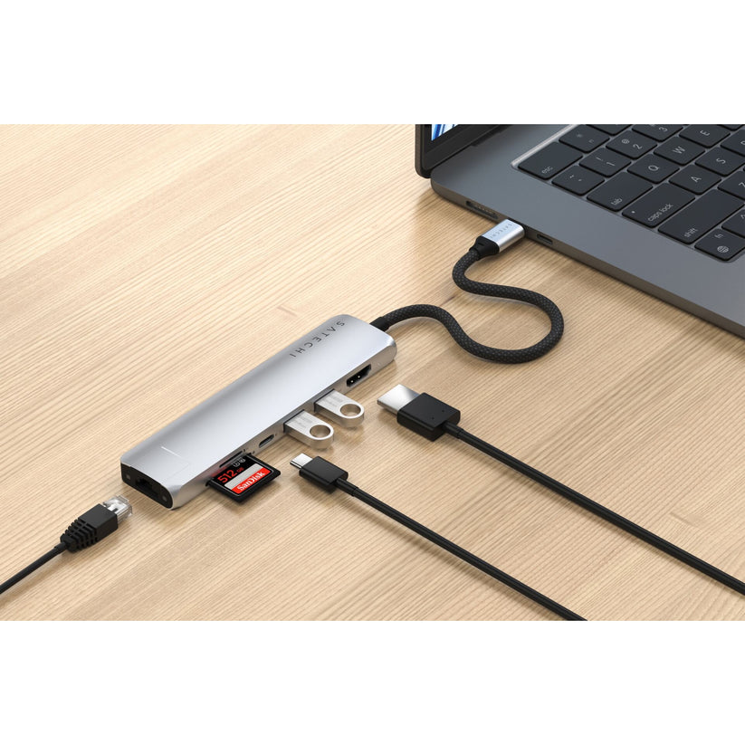 Gümüş renkli, Ethernet özellikli bir Satechi USB-C 7′si 1 arada İnce Çoklu Bağlantı Noktalı Adaptör, bir dizüstü bilgisayara bağlıdır ve USB kabloları, bir SD kart, bir microSD kart ve bir Ethernet kablosu dahil olmak üzere çeşitli cihazları barındırır; tüm bunlar açık renkli ahşap bir yüzey üzerinde düzenli bir şekilde yer almaktadır. Bu çok yönlü Satechi adaptör, çalışma alanınızı düzenli tutmanıza ve cihazlarınız arasında sorunsuz bir bağlantı kurmanıza yardımcı olur.