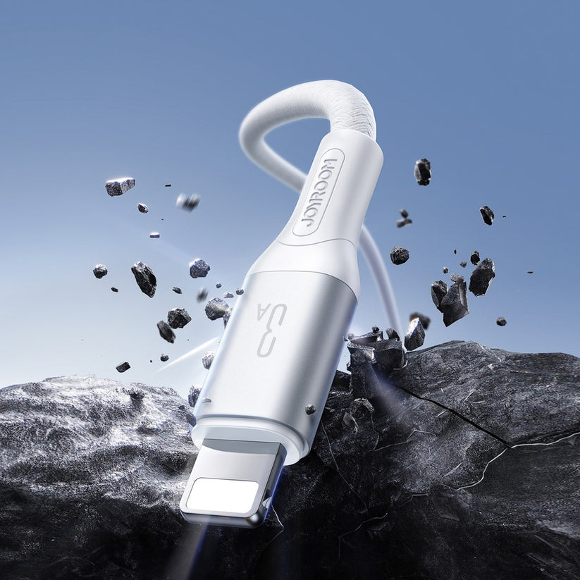 Eine Nahaufnahme des Joyroom USB-A-auf-Lightning-Kabels (1 m, 15 W, USB 2.0) in Weiß, auf dem das JOYROOM-Logo deutlich zu erkennen ist, während es kraftvoll durch schwarzen Fels dringt und dabei vor dem Hintergrund eines blauen Himmels Trümmerteile durch die Luft fliegen – als Symbol für schnelles Aufladen und zuverlässige Datenübertragung.