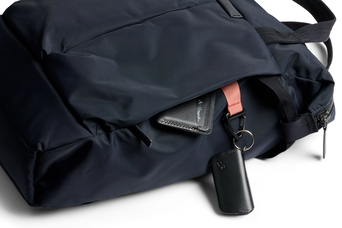 Ein kompakter Rucksack, der Bellroy Tokyo Totepack Compact in Navy, mit teilweise geöffnetem Reißverschluss, zeigt ein schwarzes Portemonnaie und einen Schlüsselanhänger mit schwarzem Anhänger und braunem Band; außerdem verfügt die Tasche über ein Laptopfach.