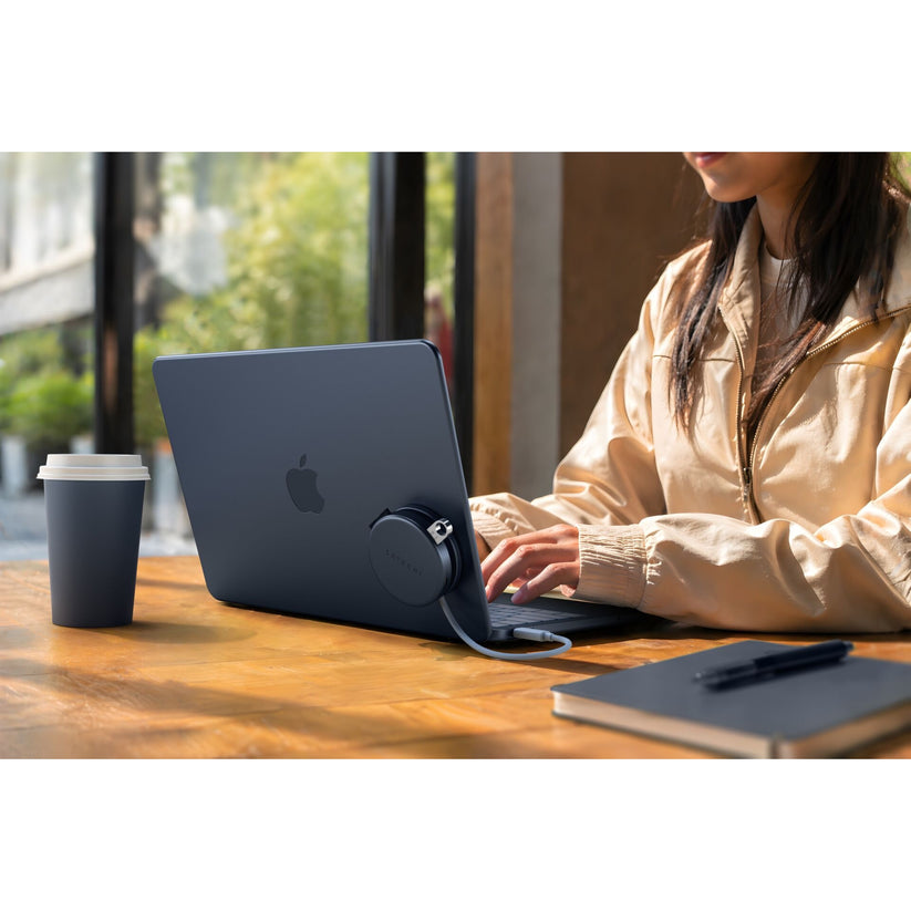 Eine Person in einer hellbraunen Jacke tippt an einem Holztisch auf einem dunklen Apple-Laptop, neben einem Notizbuch, einem Stift, einer Kaffeetasse und einem aufgerollten Ladekabel. Der Satechi OntheGo 5-in-1-Multiport-Adapter (Indigo) von Satechi liegt daneben und bietet in dem hellen, modernen Café praktische Anschlussmöglichkeiten für alle Geräte.