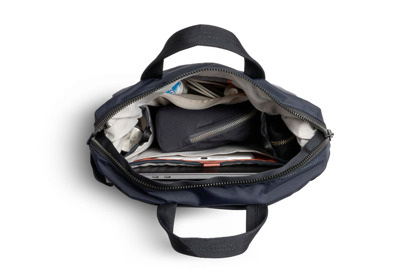 Ein von Bellroy hergestelltes „Bellroy Tokyo Totepack Compact“ in der Farbe „Navy“ ist vor einem weißen Hintergrund zu sehen: Der Reißverschluss ist geöffnet und gibt den Blick auf die ordentlich verstauten Gegenstände im Inneren frei – darunter Kleidung, ein kleines Täschchen, eine Wasserflasche sowie ein Tablet oder ein anderes Gerät, das sicher im speziellen Laptopfach verstaut ist.