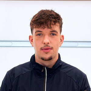 Ein junger Mann mit kurzen braunen Haaren und heller Gesichtsbehaarung, der eine dunkle Jacke mit Reißverschluss trägt, steht vor einem schlichten weißen Hintergrund mit einer schwachen horizontalen Linie hinter ihm.