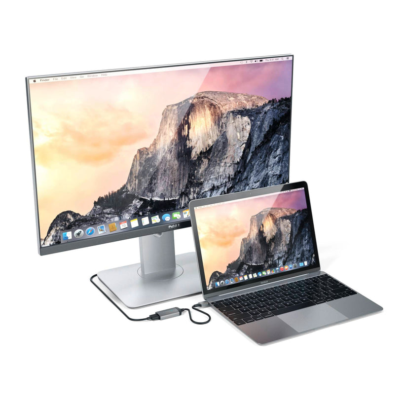 Ein Laptop, der an einen Breitbildmonitor angeschlossen ist – auf beiden Bildschirmen ist dasselbe Hintergrundbild mit einer Berglandschaft zu sehen –, steht auf einer weißen Fläche, wobei ein schwarzes Kabel die beiden Geräte über den Satechi Typ-C-zu-4K-HDMI-Adapter in Space Grau miteinander verbindet.