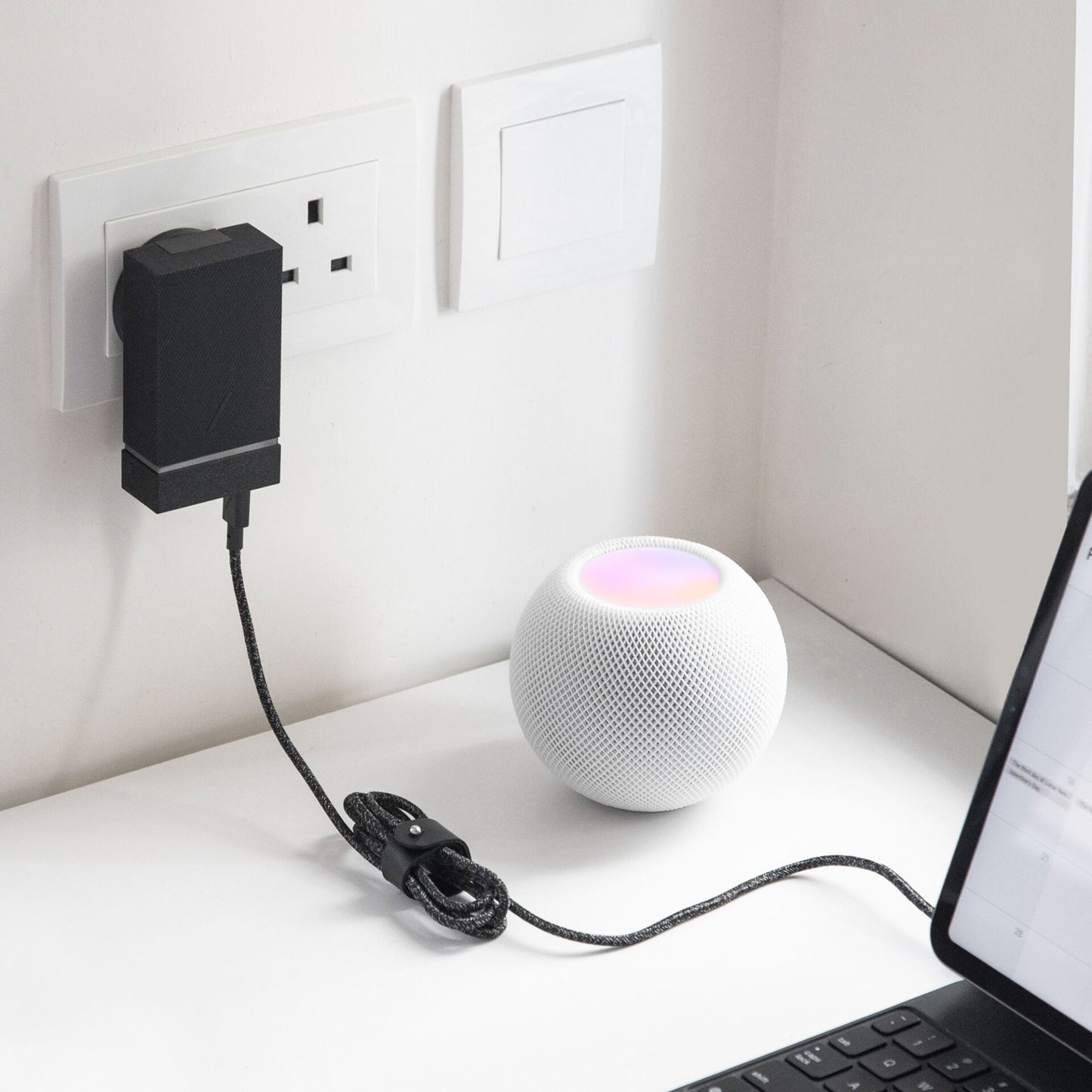 Ein Native Union-Kabel vom Typ USB-C auf USB-C (1,2 m) in der Farbe „Cosmos“ ist an eine Steckdose angeschlossen und versorgt einen kleinen weißen Smart-Lautsprecher mit leuchtender Oberseite mit Strom, der neben einem teilweise sichtbaren Laptop auf einem weißen Schreibtisch steht.