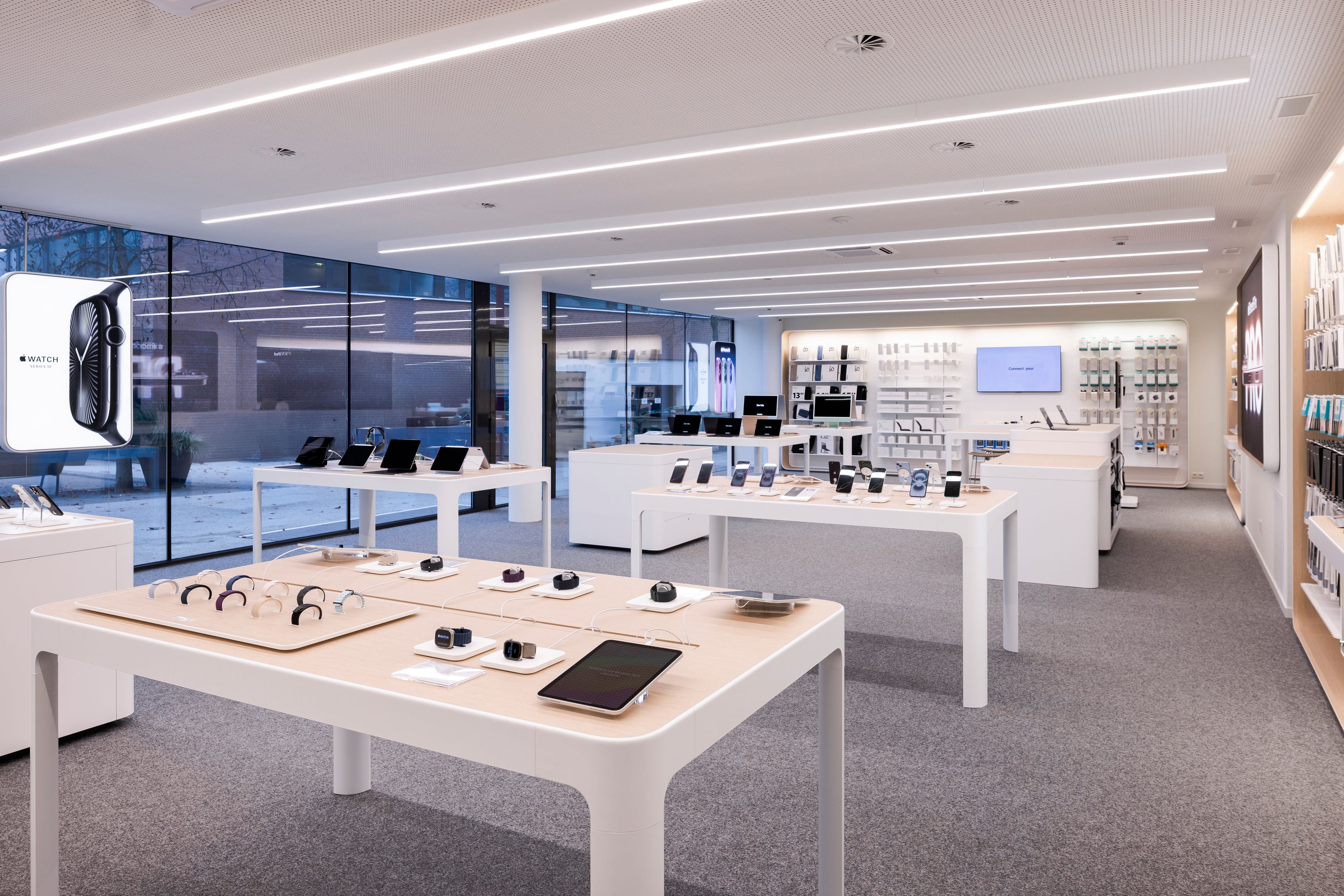 Ein modernes Elektronikgeschäft mit heller Beleuchtung und Schautischen, auf denen Apple-Smartphones, Tablets, Smartwatches und Zubehör präsentiert werden. Regale säumen die Wände, und große Glasfenster lassen natürliches Licht ins Innere.