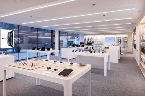 Ein modernes Elektronikgeschäft mit heller Beleuchtung und Schautischen, auf denen Apple-Smartphones, Tablets, Smartwatches und Zubehör präsentiert werden. Regale säumen die Wände, und große Glasfenster lassen natürliches Licht ins Innere.