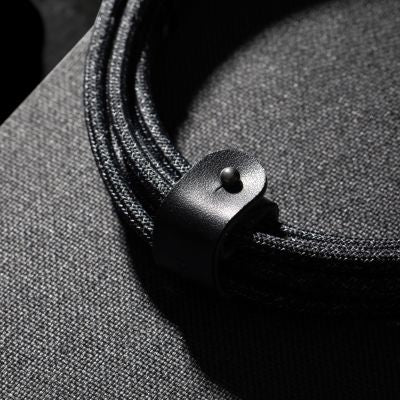 Ein gewendeltes dunkles Kabel, wie beispielsweise das „Native Union Belt Cable XL USB-C auf USB-C 3 m Cosmos“, ist mit einem schwarzen Lederriemen und einem Druckknopf befestigt und liegt auf einer strukturierten dunklen Stoffoberfläche, die von dramatischen Licht- und Schatteneffekten umspielt wird.